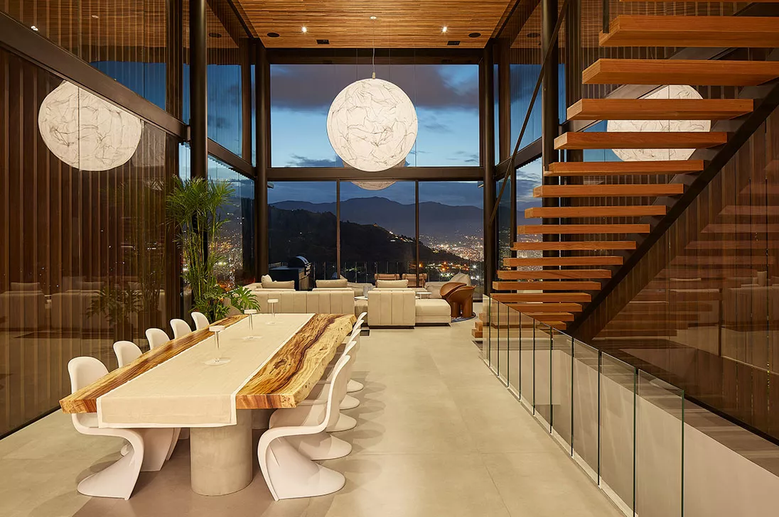 A spacious, modern dining and living area with floor-to-ceiling glass walls showcasing a stunning cityscape view at dusk. The room features warm pendant lights, a natural wood dining table, sleek white chairs, and a floating staircase, creating a blend of elegance and functionality.
