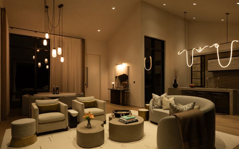 A cozy and elegant living space featuring soft ambient lighting with modern pendant lights, recessed ceiling fixtures, and a unique sculptural light over the kitchen. The room includes plush seating, round tables, and neutral tones, creating a warm and inviting atmosphere.